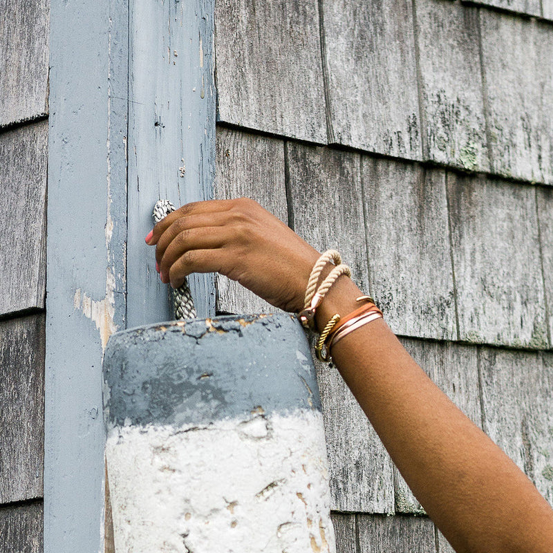 Woman placing a buoy on shingled building wearing Sailormade double wrap rope bracelet in wheat with rose gold brummel clasp and slim fid cuff in Rockport, Massachusetts.