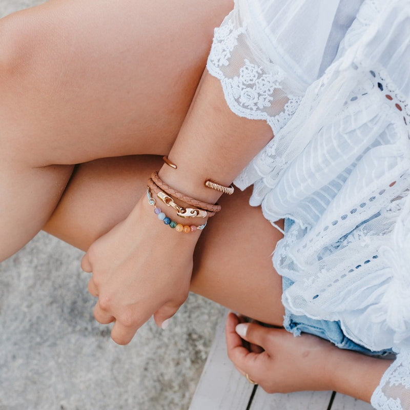 Woman by beach wearing sailormade nautical rose gold brummel bracelet with baked brown leather double wrap stacked with the slim fid cuff and rainbow rayminder uv awareness bracelet.