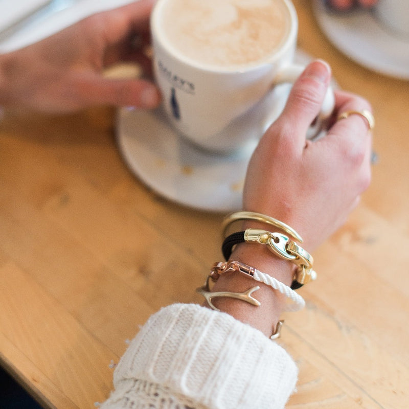 Woman wearing a casual sailormade nautical bracelet stack while drinking coffee in marblehead. Brass Fid cuff bracelet paired with rose gold lure single wrap rope brummel bracelet in white, reef cuff in satin brass, and Charter Big Brummel bracelet.