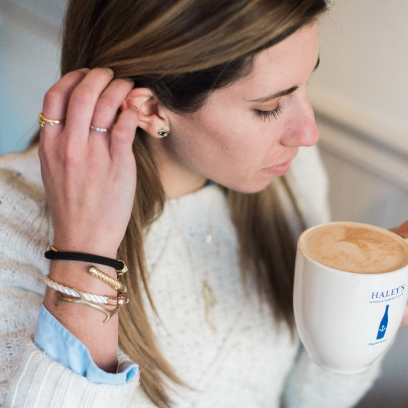 Woman wearing a casual sailormade nautical bracelet stack while drinking coffee in marblehead. Brass Fid cuff bracelet paired with rose gold lure single wrap rope brummel bracelet in white, reef cuff in satin brass, and Charter Big Brummel bracelet.